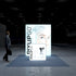 A frontal view of the Pixlip GO booth highlighting its illuminated backdrop with a striking portrait of a person with light hair and vibrant blue eyes. The compact counter in the foreground displays transparent bottle designs. A person walks past the booth carrying a bag.